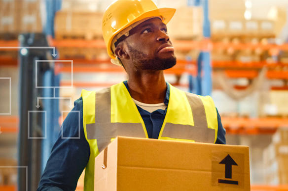 Man wearing hard helmet and high visibility vest holding a box standing in a warehouse