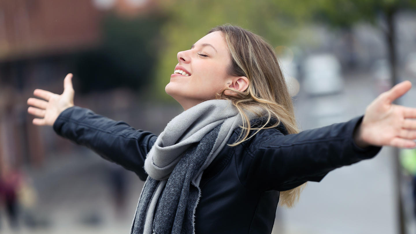 Smiling young woman breathing fresh air and raising arms in the city.