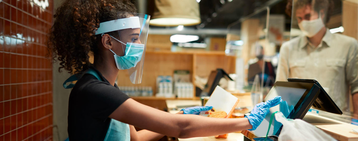 a cashier wearing a face shield and mask rings up a customer's food order