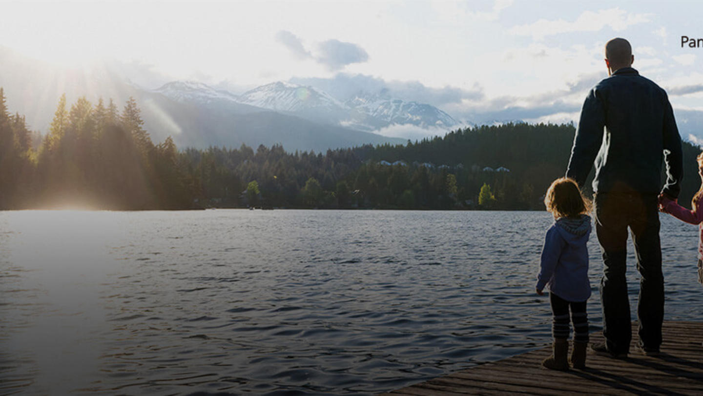 A father stands on a dock in a lake, staring at the mountains and pine trees with his children