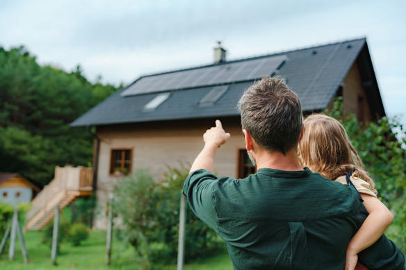 Rear view of dad holding her little girl in arms and showing at their house with installed solar panels. Alternative energy, saving resources and sustainable lifestyle concept.