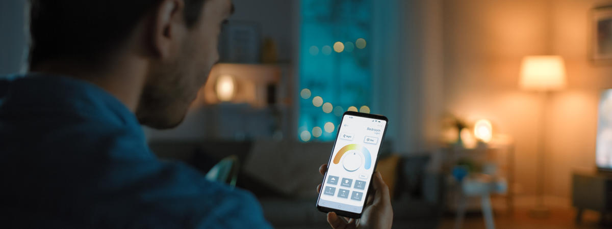 Young Handsome Man Gives a Voice Command to a Smart Home Application on His Smartphone To Change Light Temperature and Set a Comfortable Lighting. It's a Cozy Evening in Living Room.